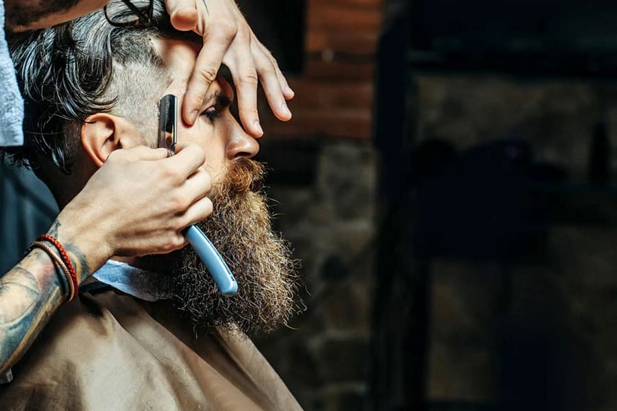 un homme à barbe est sous la main d'un barbier qui égalisé la coupe